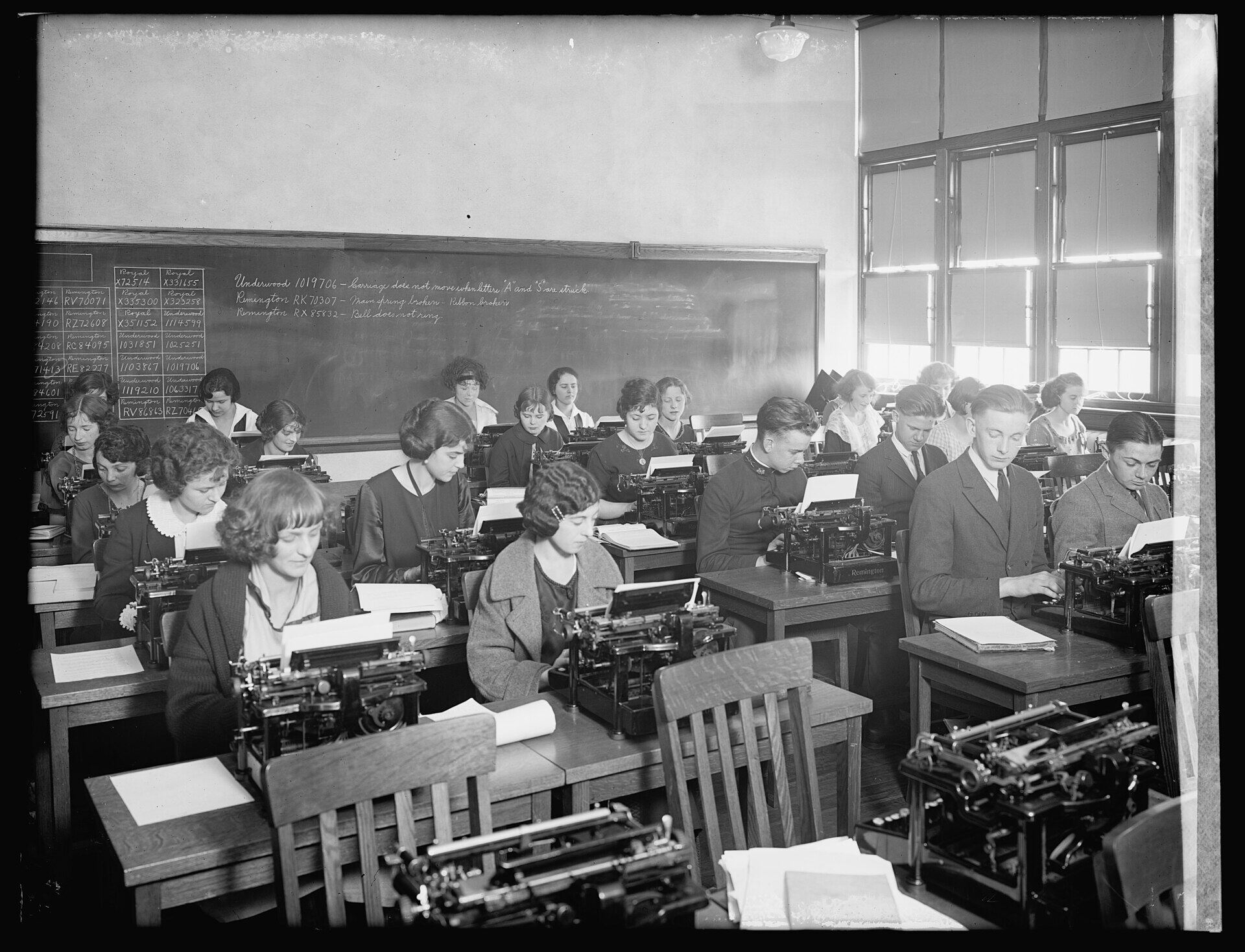 Eine Schulklasse übt an Schreibmaschinen, vorne an der Tafel stehen Übungszeilen